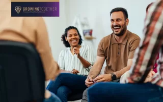 Adult men and women sit in a circle for group therapy while smiling and laughing.