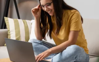 A young woman wearing glasses smiling while looking at her laptop.