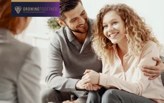 A smiling couple sits closely together, holding hands during a therapy session, while speaking with a therapist who is partially visible in the foreground.