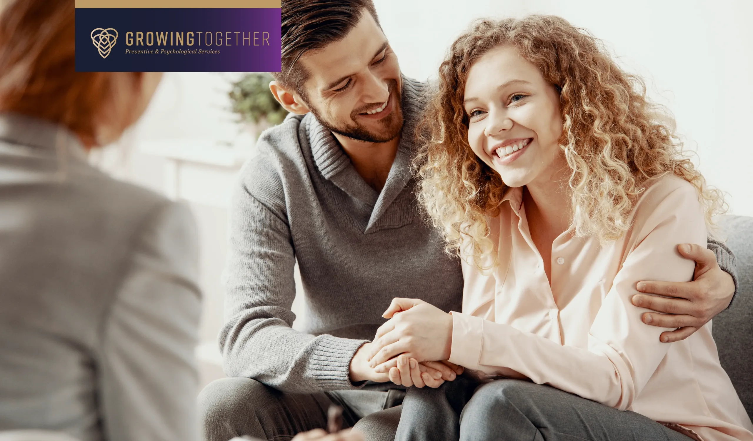 A smiling couple sits closely together, holding hands during a therapy session, while speaking with a therapist who is partially visible in the foreground.